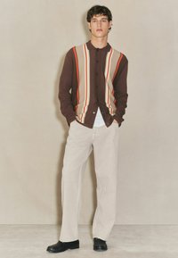 Brauner, langärmliger Strickcardigan mit mehrfarbigen vertikalen Streifen und Knopfverschluss. Kombiniert mit hellbeigen, weiten Hosen und schwarzen Schuhen.