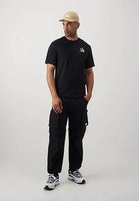 Hombre con una gorra beige, camiseta de manga corta negra, pantalones cargo negros y zapatillas blancas de pie contra un fondo liso.