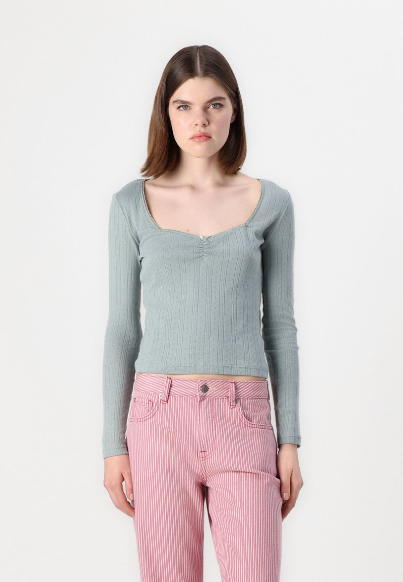 Pitkähihainen, vaaleanvihreä paita, jossa on pyöreä pääntie, ribbikuvio ja keskellä rypytys, yhdistettynä vaaleanpunaisiin ja valkoisiin raidallisiin housuihin.