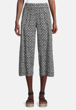 Person trägt schwarz-weiß gemusterte, weit geschnittene und kurz geschnittene Hosen sowie schwarze Plateausandalen mit Knöchelriemen.