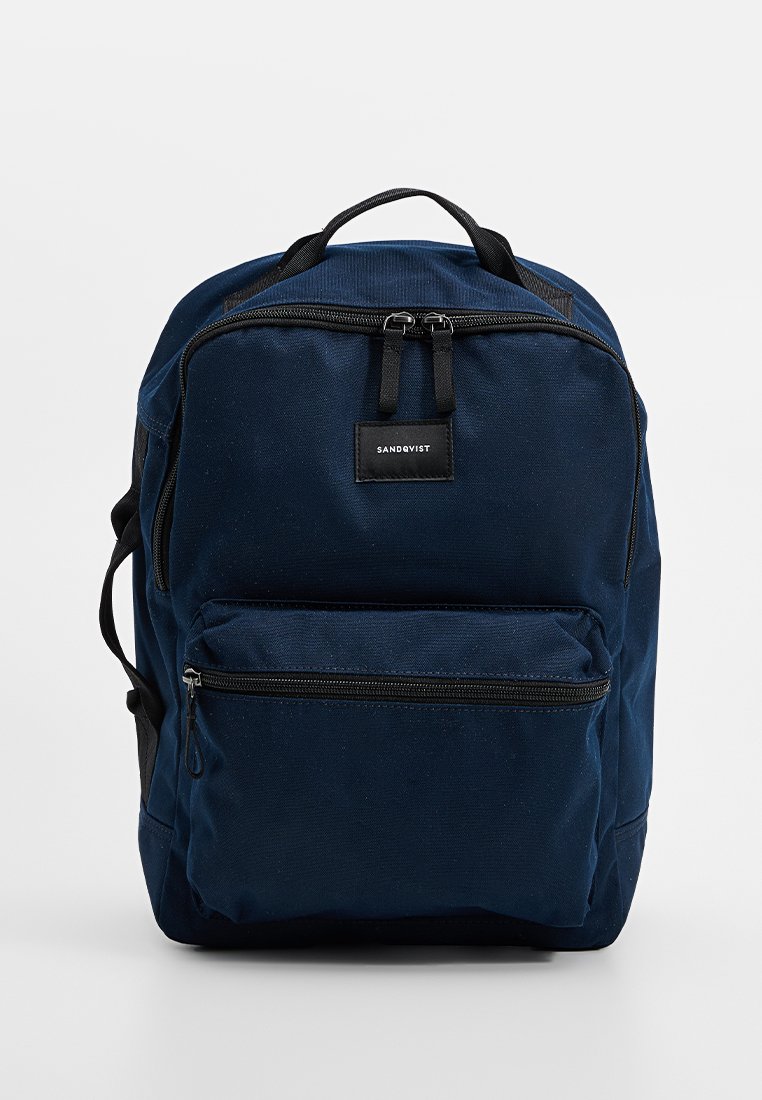 Mochila de lona azul marino con un bolsillo frontal con cremallera, asa superior y correas de hombro ajustables. Cuenta con una etiqueta de marca negra en la parte frontal.