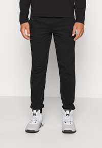 Musta urheiluhaalari, joka on valmistettu sileästä materiaalista, jossa on kapeneva muotoilu, joustavat hihnat ja mukava istuvuus. Yhdistetty harmaisiin tennareihin.