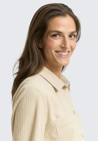 Beżowa koszula z sztruksu o luźnym kroju, z kołnierzykiem, guzikami i kieszonką na piersi. Materiał ma pionowe prążki i miękką fakturę.