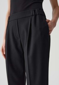 Pantaloni neri su misura con una texture liscia, caratterizzati da una vita alta, pince e tasche laterali. Il materiale sembra leggero e formale.