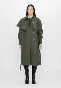 Σκούρο πράσινο αδιάβροχο παλτό με κολάρο σε στυλ κάπας, κουμπιά κλεισίματος, ρυθμιζόμενα μανίκια και ευθεία γραμμή. Φοριέται πάνω από μαύρες μπότες.