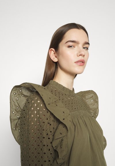 Blouse vert olive avec des motifs découpés complexes, épaules volantes et corsage froncé. Fabriquée en tissu léger avec des détails texturés.
