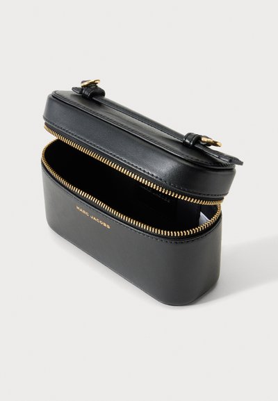 Ορθογώνια μαύρη θήκη Marc Jacobs με χρυσό φερμουάρ μερικώς ανοιχτό, μαύρη δερμάτινη λαβή στην κορυφή, απλό ανοιχτό φόντο.