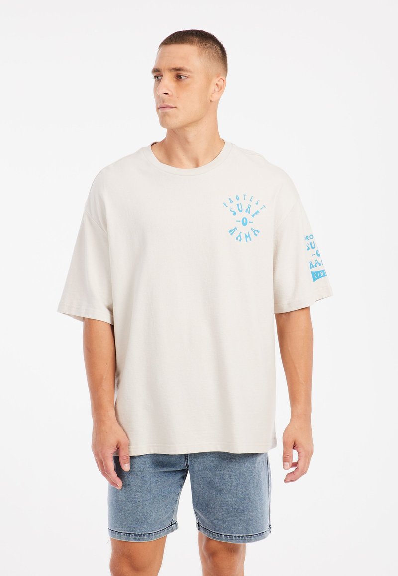 Světlé béžové oversized tričko vyrobené z měkké bavlny. Na hrudi a na rukávu jsou modré grafické nápisy. Krátké rukávy a kulatý výstřih.
