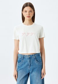 Witte cropped t-shirt met roze lint borduursel op de borst, met korte mouwen en een ronde halslijn, gecombineerd met blauwe denim jeans.
