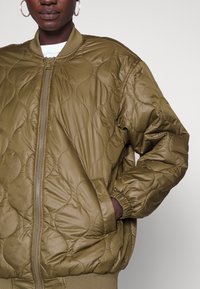 Oliwkowo-zielona pikowana kurtka bomberka z zamkiem błyskawicznym z przodu, ściągaczami na rękawach i zaokrąglonym kołnierzem. Ma jedwabistą fakturę oraz boczne kieszenie.