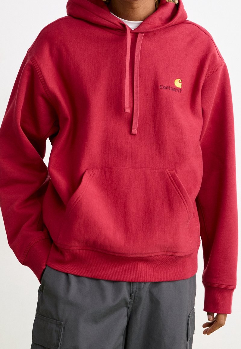 Personne portant un sweat à capuche rouge Carhartt avec poche avant et pantalon cargo gris, debout avec une main dans la poche et l'autre détendue.