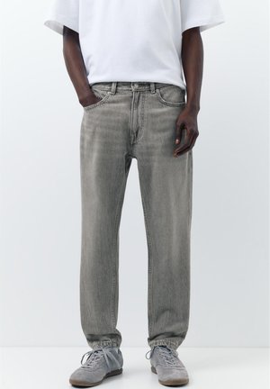 Mann trägt ausgewaschene graue Denim-Jeans mit elastischen Bündchen, graue Sneakers und ein weißes kurzärmeliges Hemd, steht mit einer Hand in der Tasche.