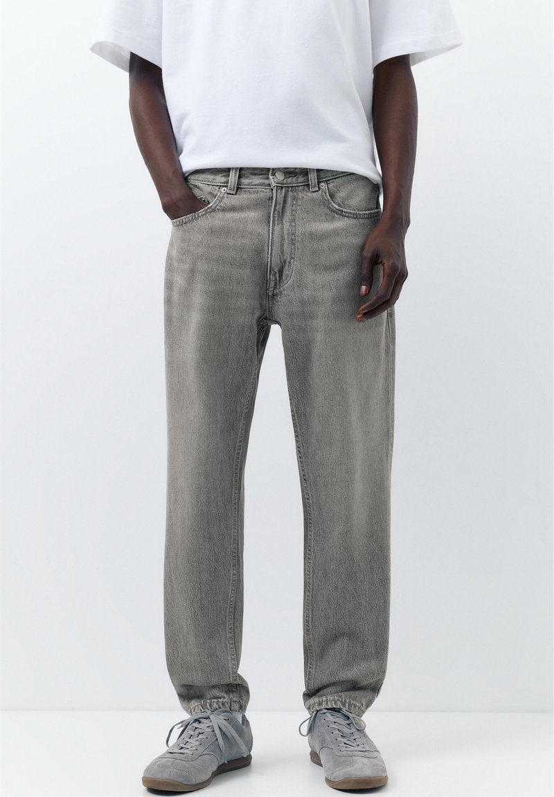 Homme portant un jean en denim gris délavé avec des poignets élastiques, des baskets grises et une chemise blanche à manches courtes, debout avec une main dans la poche.
