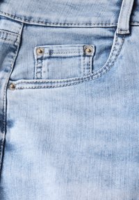 Vaaleansininen farkkukangas, jossa on teksturoitu pinta. Siinä on pieni etutasku metallinapeilla, hienovarainen kuluminen ja ommeltuja saumoja.