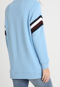 Persona che indossa una felpa azzurro chiaro a maniche lunghe con strisce nere, bianche e rosso scuro sui polsini, abbinata a jeans blu.