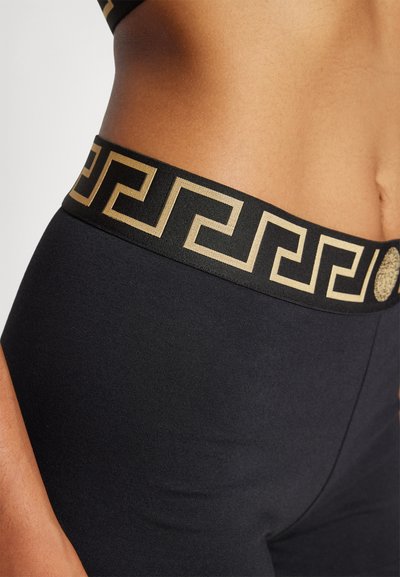 Svarta högmidjade leggings med ett guldgrekiskt nyckelmönster på den elastiska midjebandet, med en jämn och elastisk tygtextur.