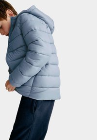 Pojke i ljusblå pufferjacka med huva, justerar fållen, står mot en enfärgad vit bakgrund.