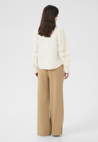 Blusa di crema con maniche a sbuffo e dettagli ricamati, abbinata a pantaloni beige a vita alta e a gamba larga, in tessuto morbido e con una vestibilità rilassata.