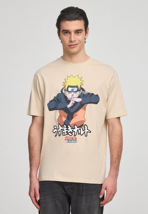 T-shirt in cotone beige con una grafica di un personaggio con capelli gialli a punta, vestito con un completo nero e arancione, accompagnato da un testo stilizzato sotto.
