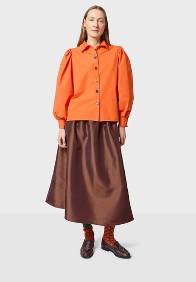 Oranssi puuvillapaita, jossa on puffihihat, värikkäät napit ja ruskea satiininen pitkä hame. Ruskeat kengät ja kuvioidut oranssit sukat täydentävät asun.