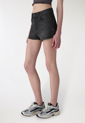 Persona che indossa pantaloncini di jeans neri e scarpe da ginnastica argentate, in piedi contro uno sfondo chiaro e uniforme.