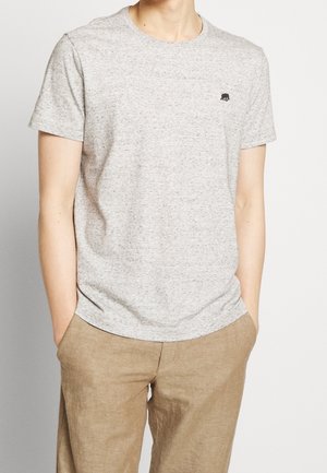 Mann trägt ein hellgraues, kurzärmeliges T-Shirt mit kleinem schwarzem Logo auf der Brust und beige Hose mit Händen in den Taschen.