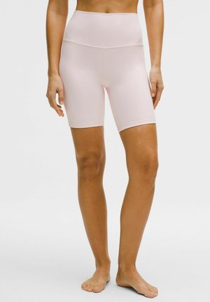 Kvinde iført lyserøde cykelshorts med høj talje, stående barfodet mod en ensfarvet hvid baggrund.