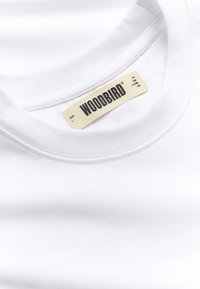 Λευκό T-shirt με στρογγυλή λαιμόκοψη και μια μπεζ ετικέτα ραμμένη μέσα στη λαιμόκοψη που εμφανίζει το "WOODBIRD" με έντονα μαύρα γράμματα και λεπτομέρειες μεγέθους.