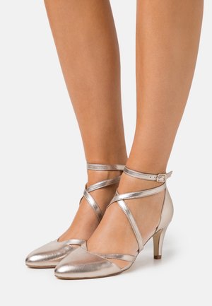 Zapatos de tacón metálicos con correas en plata, con punta afilada, tacón bajo y correas cruzadas en el tobillo. Textura suave y detalles de diseño elegantes.