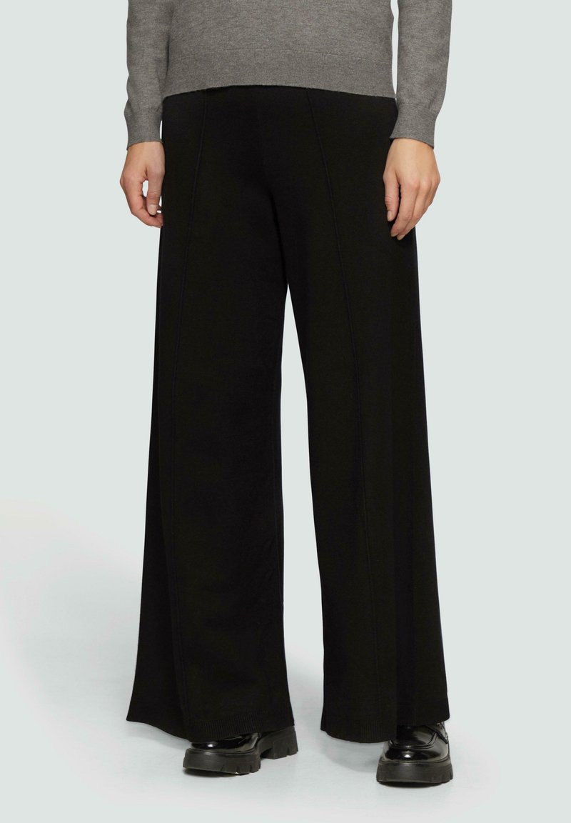 Zwarte wijde broek gemaakt van een zachte stof, met naaddetails en een hoge taille. Gecombineerd met een grijze trui en zwarte plateauschoenen.