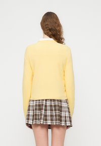 Pull en maille jaune à manches longues, associé à une jupe à carreaux marron et crème. Le pull présente un col rond et un ourlet côtelé.
