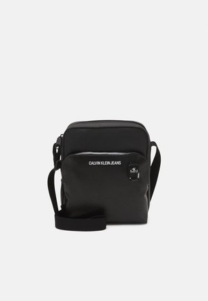 Sac bandoulière noir Calvin Klein Jeans avec poche zippée à l'avant, sangle d'épaule réglable et étiquette de marque sur fond uni.