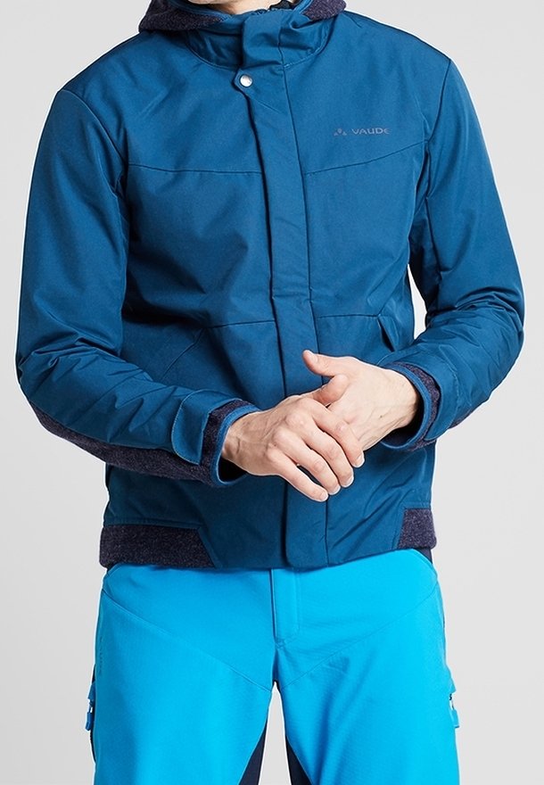 Homme portant une veste Vaude bleue avec des accents foncés et un pantalon bleu vif, les mains jointes à la taille, faisant face vers l'avant.