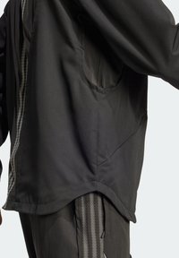 Black athletic jacket with a textured, honeycomb pattern, featuring a loose fit, side pocket, and a curved hem for mobility.