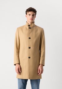 Palton din lână de culoare cămilă, cu guler înalt, șase butoane negre și buzunare laterale. Design drept, textură netedă și lungime medie.