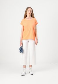 Orange kortermet skjorte, rund hals, løs passform, parret med vide hvite bukser og høye hvite joggesko, holder en blå clutch.