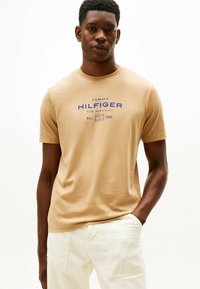 Beige bomulds T-shirt med et blåt grafisk tryk med teksten "TOMMY HILFIGER NEW YORK CITY" og "Est. 1985"; rund halsudskæring, korte ærmer.