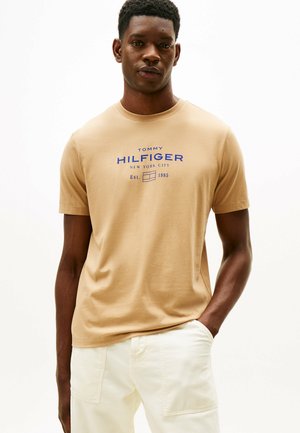Beżowy bawełnowy T-shirt z niebieskim nadrukiem graficznym, na którym znajduje się tekst "TOMMY HILFIGER NEW YORK CITY" oraz "Est. 1985"; okrągły dekolt, krótkie rękawy.