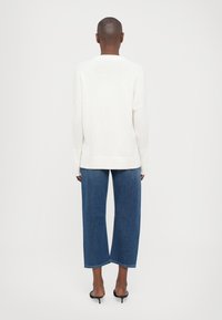 Pull en maille blanche à la coupe décontractée, avec des épaules tombantes et des accents côtelés, associé à un jean en denim bleu à jambes larges et des talons noirs.