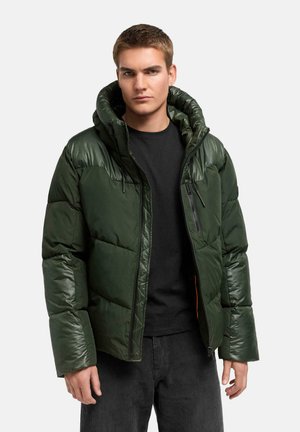 Jonge man met een donkergroen gewatteerd jack met capuchon, zwart shirt en zwarte broek, staand tegen een egale lichte achtergrond.