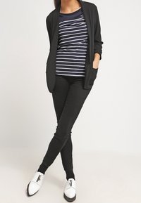 Person wearing black blazer, black skinny pants, navy blue and white striped shirt, and white shoes with zippers, standing with hands in pockets.