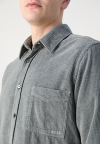 Homme portant une chemise en velours côtelé grise à boutons avec une poche poitrine brodée du logo "BOSS", visible des épaules aux lèvres.