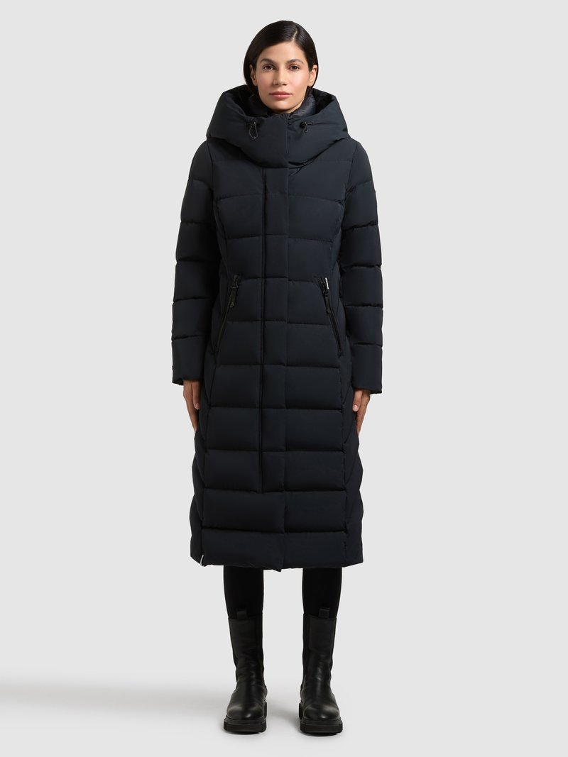 Mörkblå vadderad jacka med huva, som har en lång, rektangulär form, fickor med dragkedja och ett quiltat mönster. Komplett med svarta stövlar.