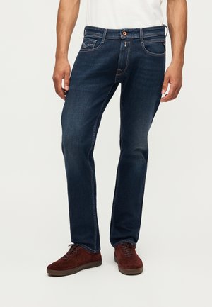 Mann, der dunkelblaue Jeans mit geradem Bein und braune Wildleder-Schnürschuhe trägt, steht vor einem schlichten hellen Hintergrund.