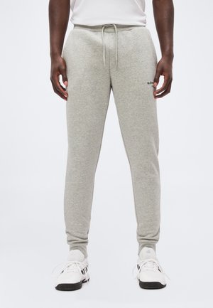 Man die lichtgrijze joggingbroek met trekkoord en witte sneakers draagt, staand tegen een effen witte achtergrond.
