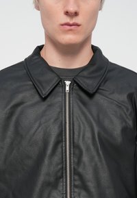 Black leather jacket with a textured finish, featuring a pointed collar and silver zipper closure, worn over a gray shirt.