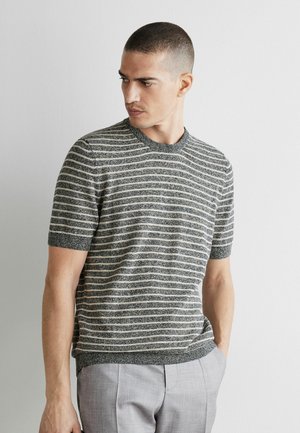 Mand med kort hår iført en grå og hvid stribet kortærmet sweater og lysegrå bukser, der kigger til siden mod en ensfarvet væg.