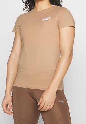 Osoba ubrana w dopasowany beżowy t-shirt Puma i brązowe legginsy Puma, stojąca z ramionami swobodnie opuszczonymi wzdłuż ciała na jednolitym tle.