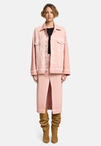 Veste en denim rose avec deux poches poitrine, associée à une jupe rose à fente avant. Le modèle porte des bottes en daim beige à talon large.