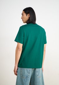 Žalia medvilnė crewneck tipo marškinėliai su trumpomis rankovėmis, pasižymintys laisvu siluetu ir glotnia tekstūra, dėvimi su šviesiai mėlynomis džinsinėmis šortais.